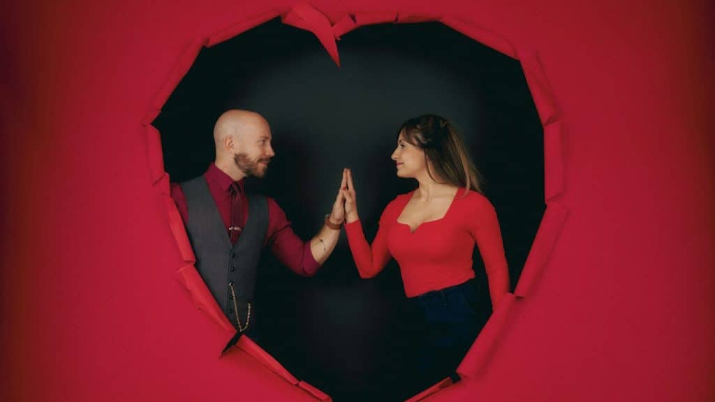 A couple standing in a red big heart are touching their hands.