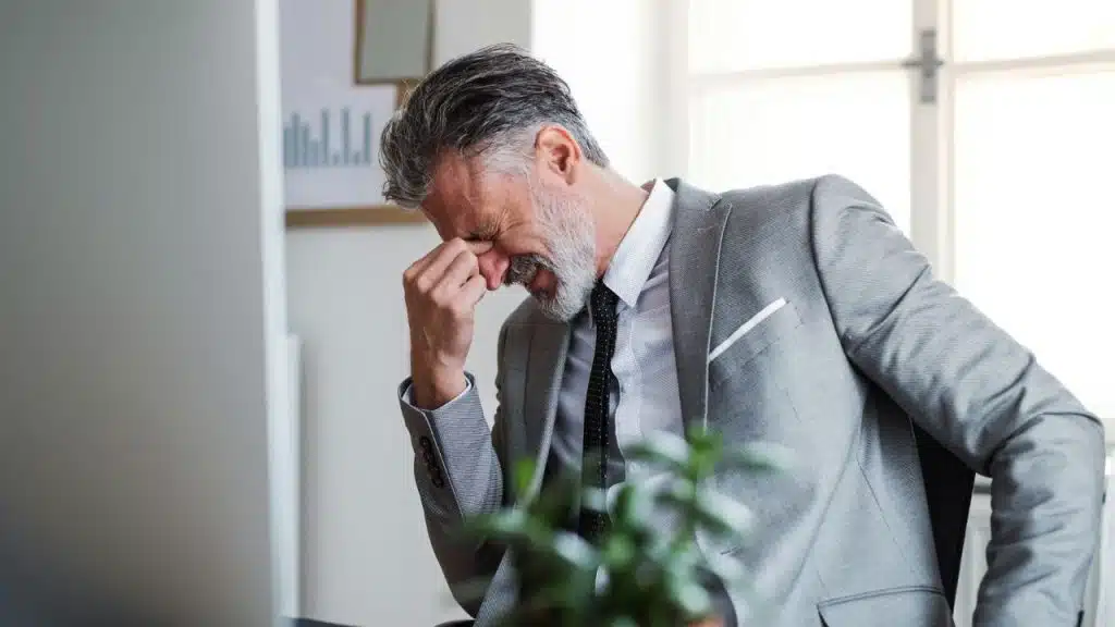 A man in a gray suit sits at a desk, pinching the bridge of his nose.