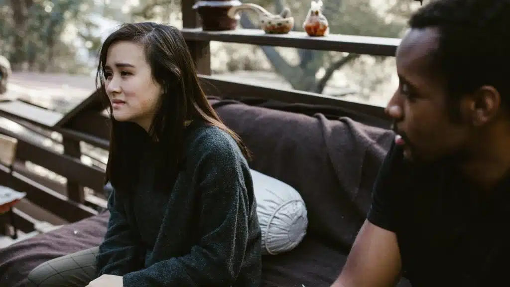 A woman with a sad expression sits outdoors on a bench next to a seated man.