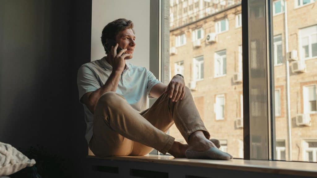 A person sitting by a window, smiling while talking on the phone.
