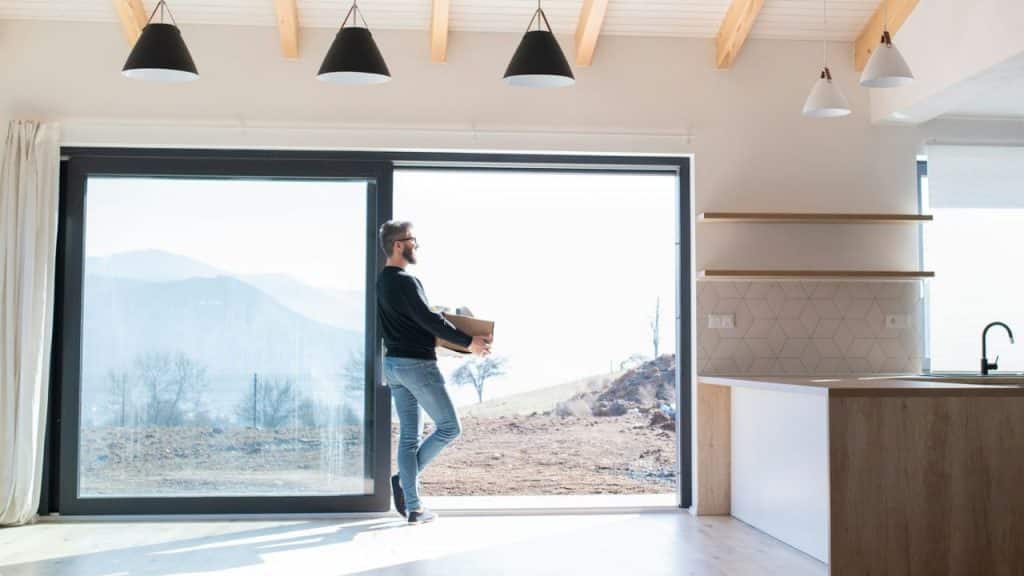 A man holds a cardboard box while looking out a large glass door in a house.