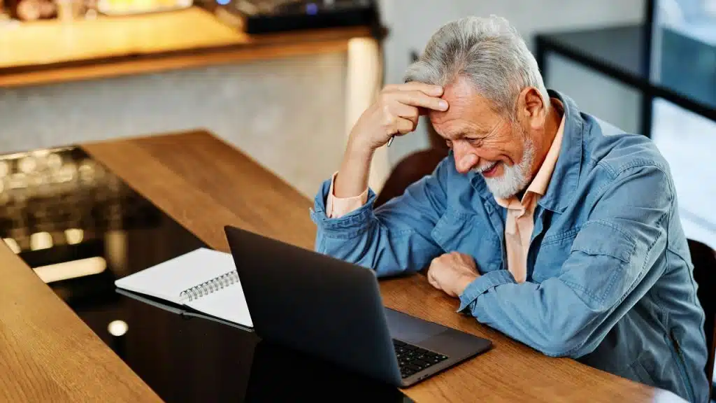 A gray-haired man smiles while looking at a laptop at a wooden desk.