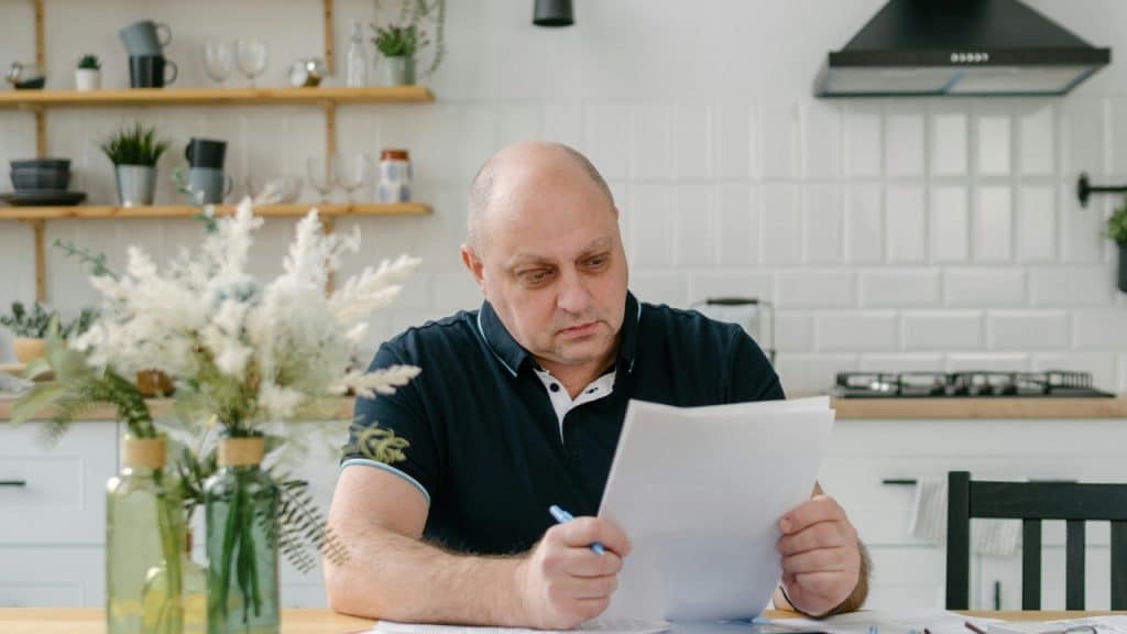 A man sits at a kitchen table looking at papers with a pen in his hand.