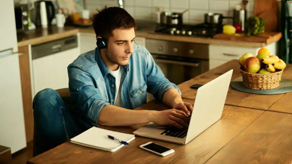 A man wearing headphones types on a laptop at a wooden table in a kitchen setting.