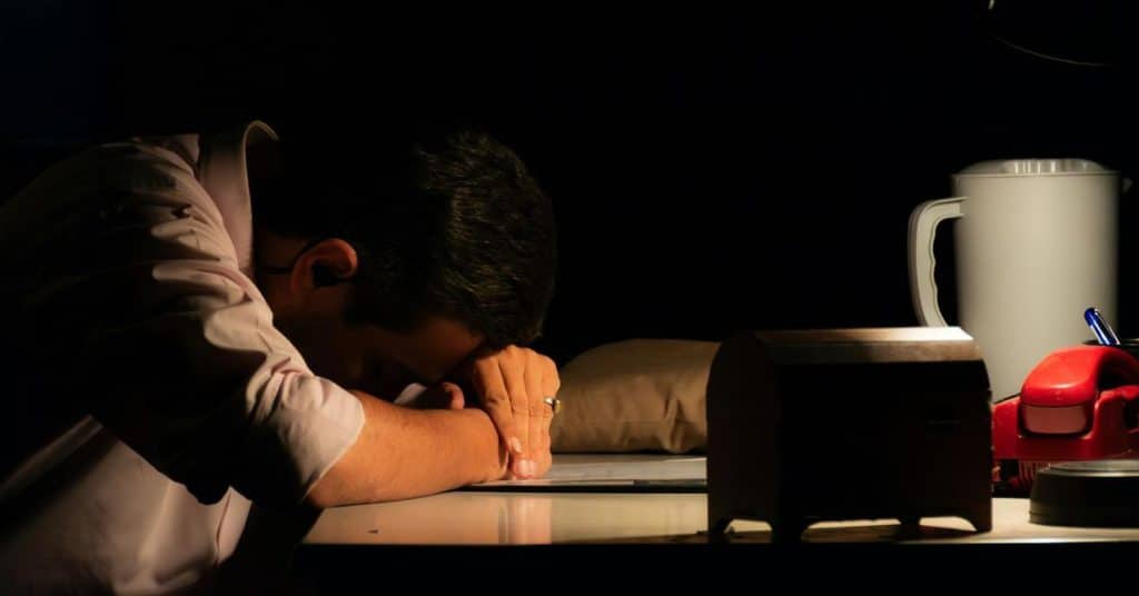 A man resting his head on a desk under dim light.