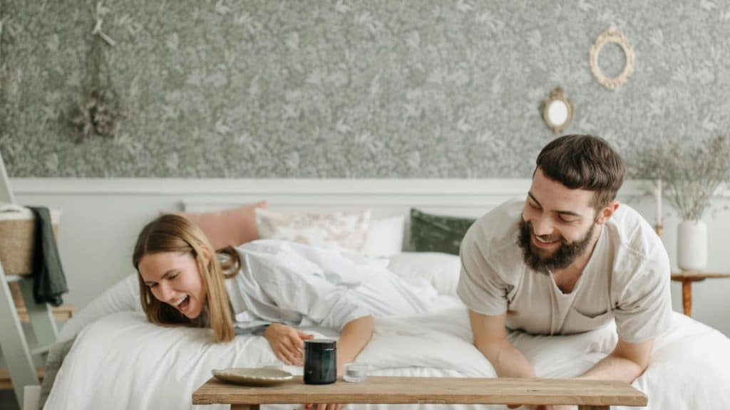A couple laughing together while leaning over a low table in a cozy bedroom.
