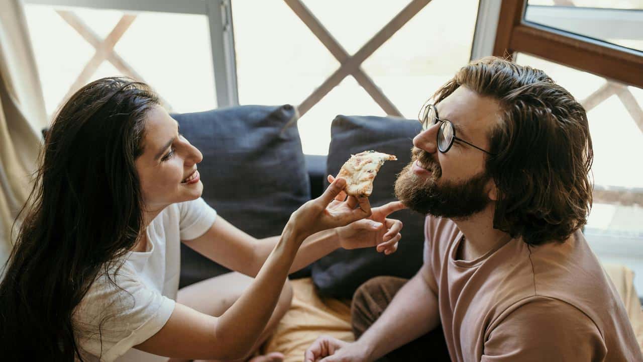 A woman smiling while feeding a man a slice of pizza at home.