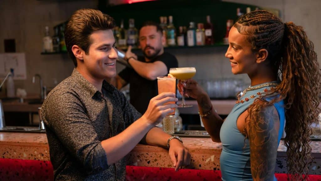 A man and a woman smiling while clinking cocktail glasses together over a pink speckled bar.