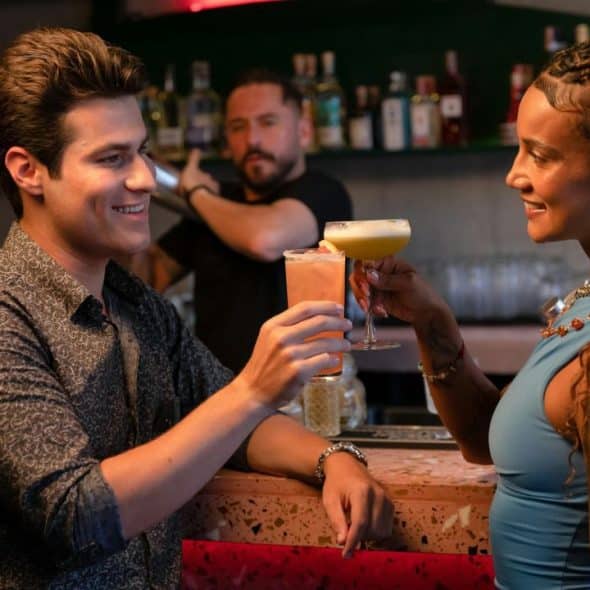 A man and a woman smiling while clinking cocktail glasses together over a pink speckled bar.