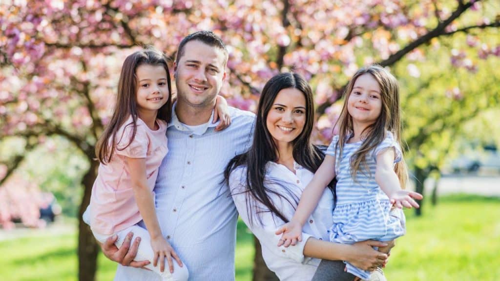 A man and woman smile while holding two young girls in front of flowering trees.