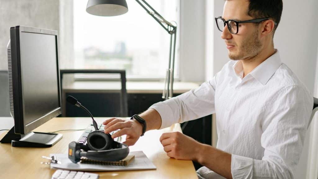 A man wearing glasses working at a desk with a computer and headset.