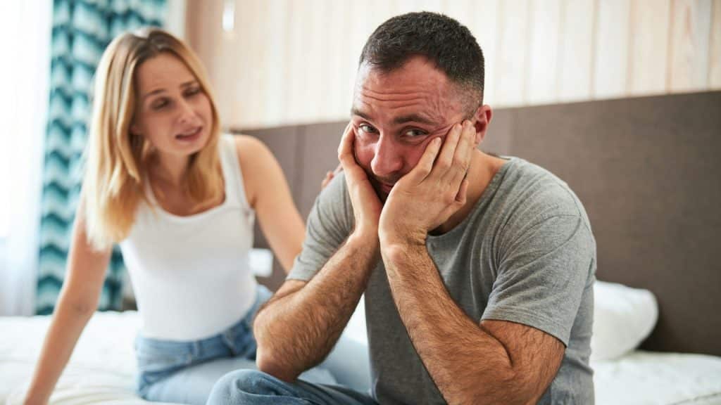 A man sits on a bed holding his face while a woman speaks behind him.