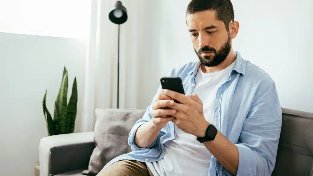 A bearded man in a blue button-down shirt sits on a sofa looking at his smartphone.