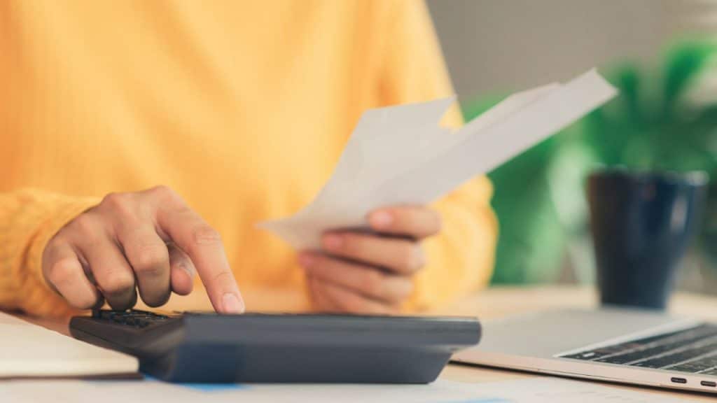 A person in a yellow sweater uses a calculator while holding several white paper slips.