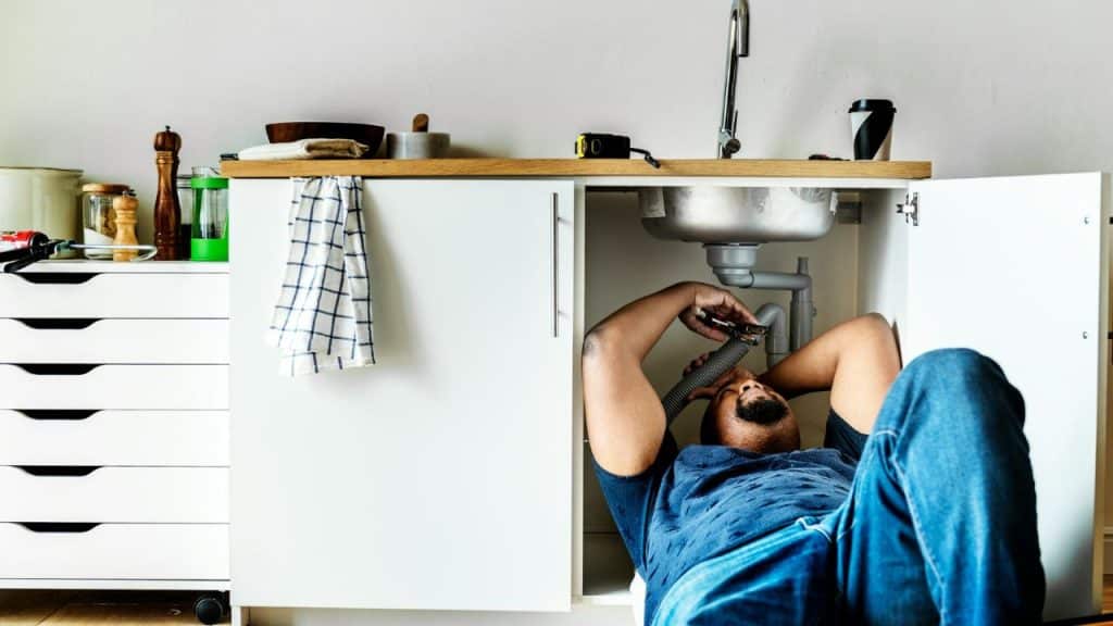 A man lies on his back on the floor while repairing pipes under a kitchen sink.