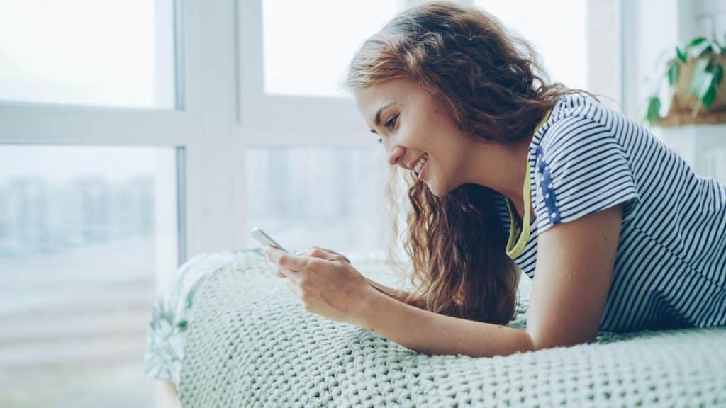 A woman smiling while using her phone on a bed near a window.