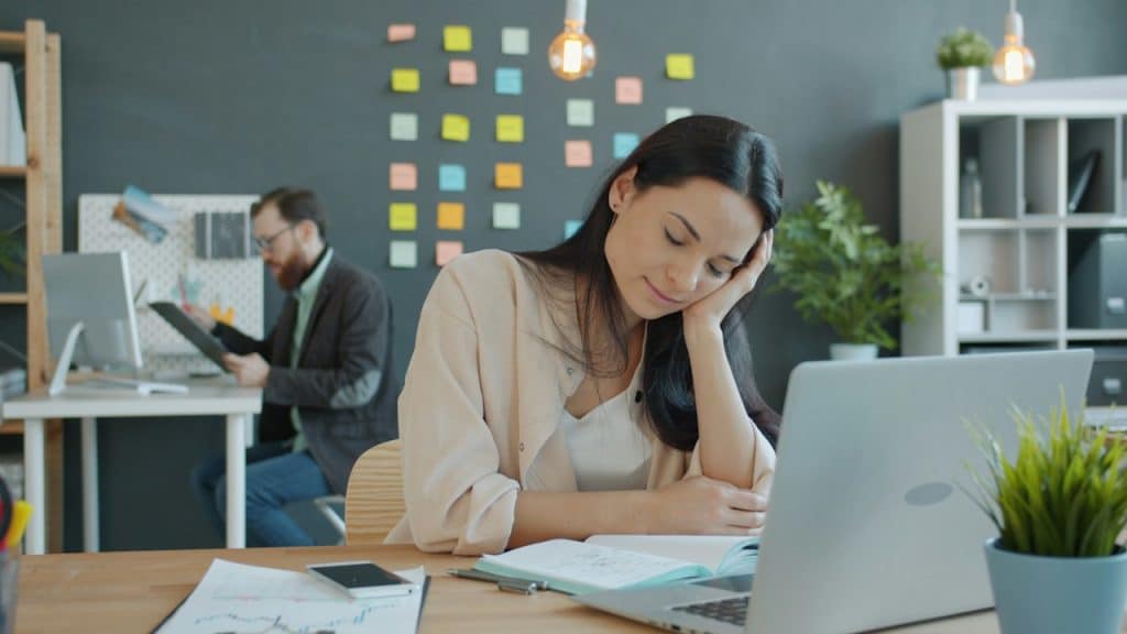 An exhausted woman sitting at a table with a laptop and a phone with a man working in the background.