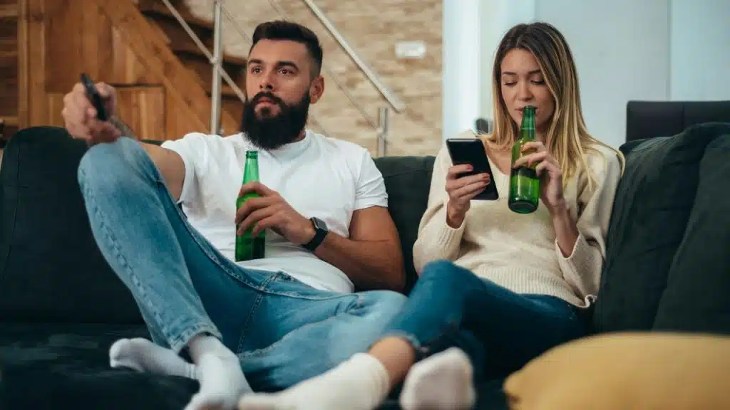 A man uses a remote while a woman looks at her phone; both hold green bottles.