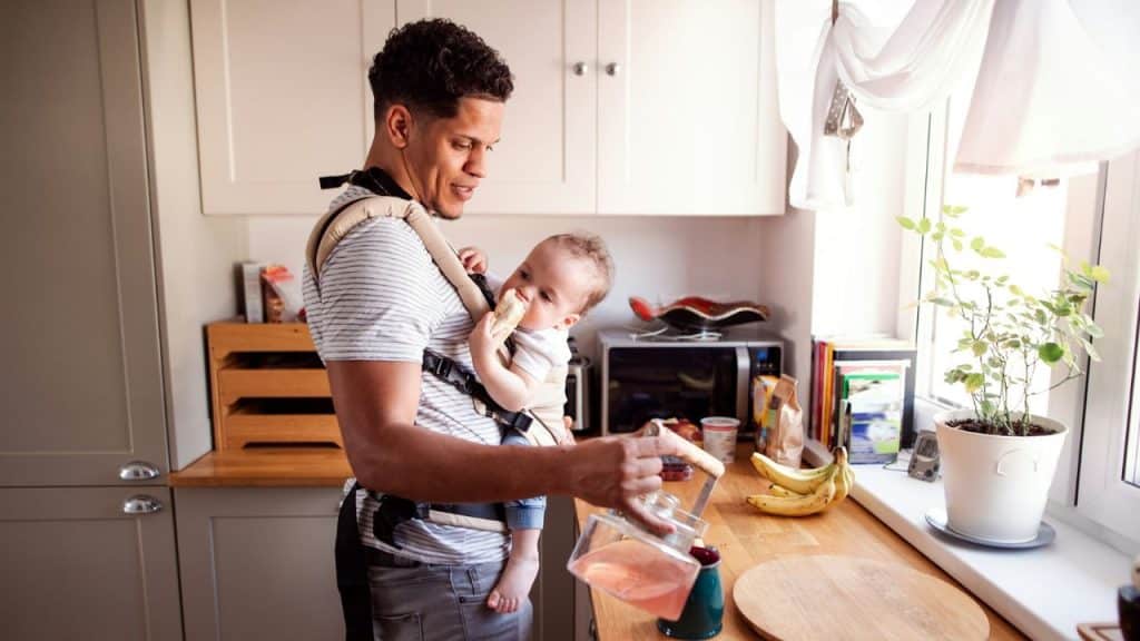 A man wears a baby carrier in a kitchen while pouring liquid into a blue mug.
