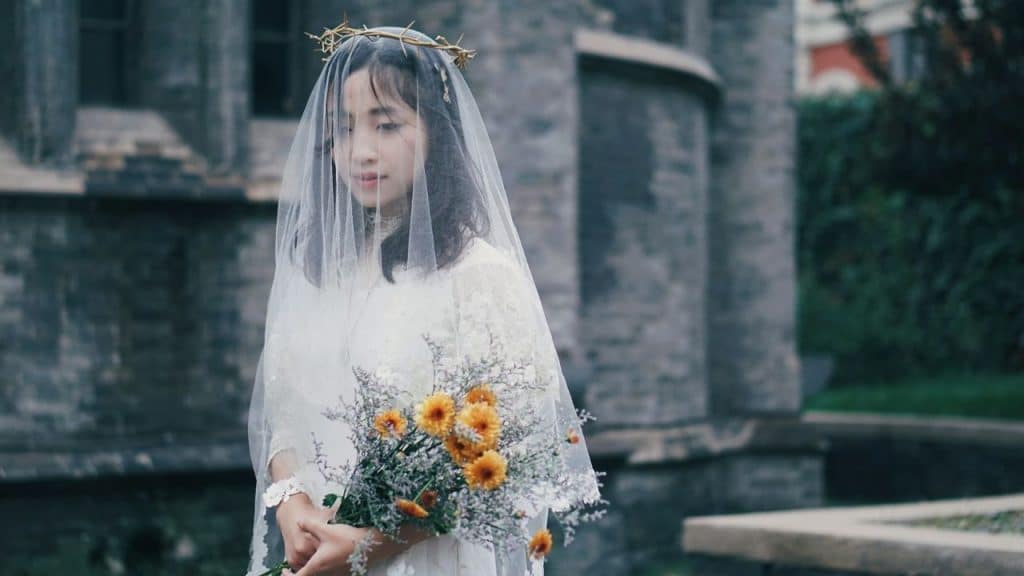 A woman in a white lace dress and veil wearing a crown of thorns holding flowers.