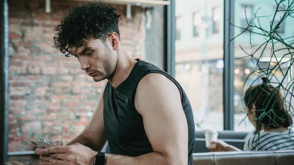 A man with curly hair looks down at a smartphone while sitting in a cafe.