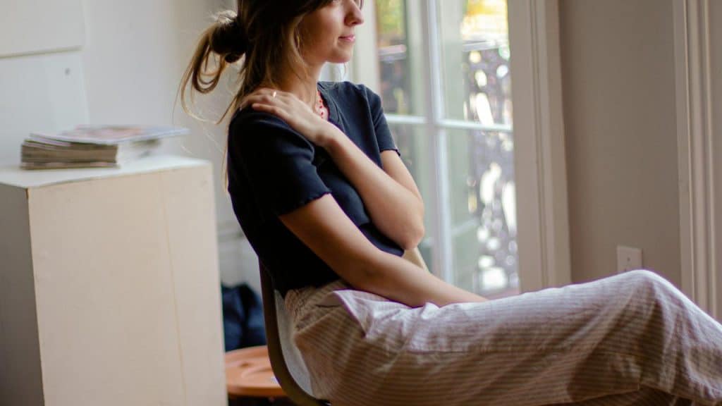 A woman sits by a window with one hand resting on her shoulder.