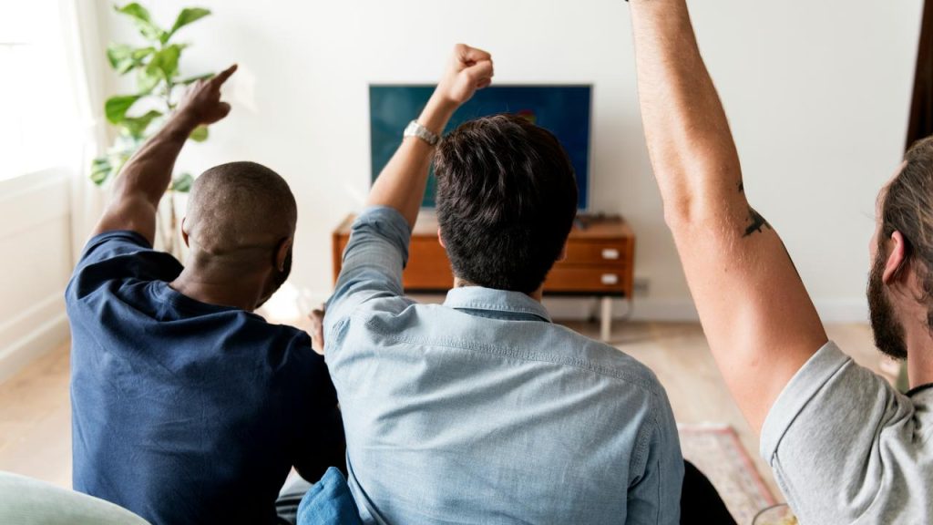 A group of people sitting on a couch with arms raised while watching a TV.