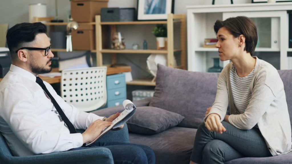 A woman sits on a sofa and speaks to a man who is holding a white clipboard.