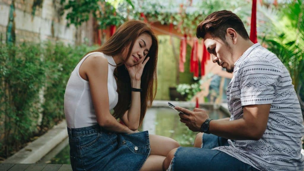 A man looks at a smartphone while a woman rests her head on her hand.
