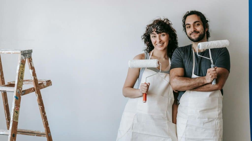 A smiling man and woman in aprons hold paint rollers in front of a wall.