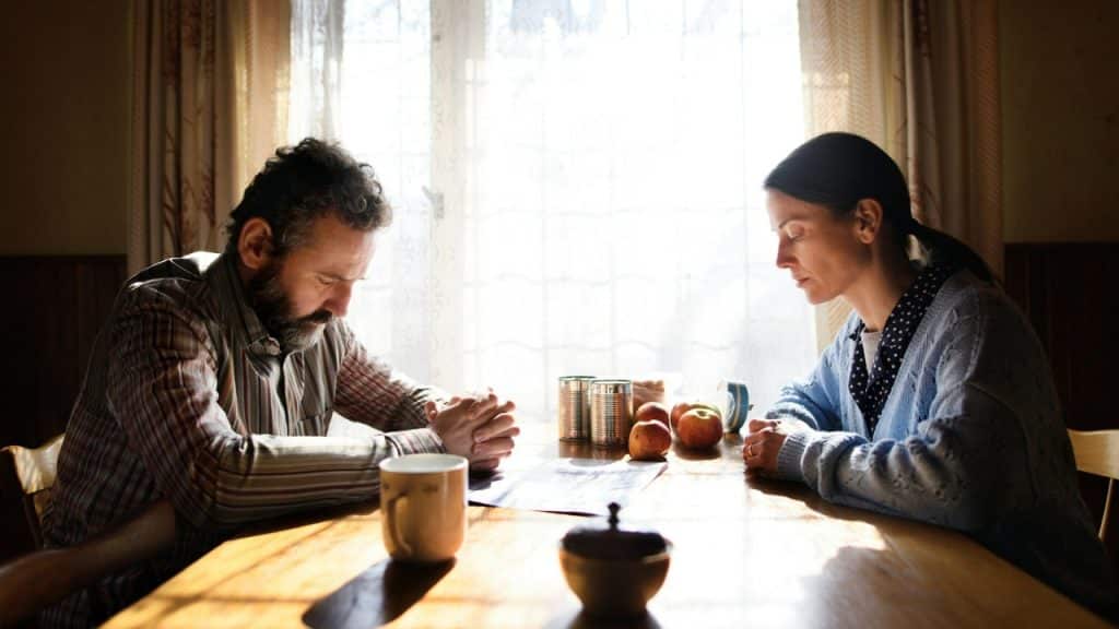 A man and woman sit at a wooden table with heads bowed near a window.