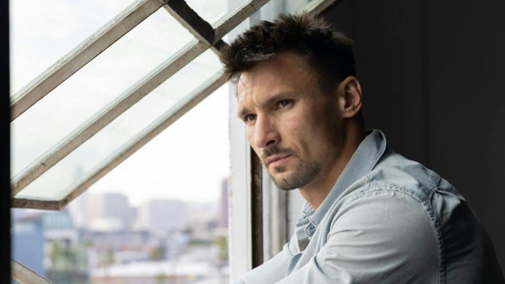A man with short dark hair looks out of a window toward a city skyline.