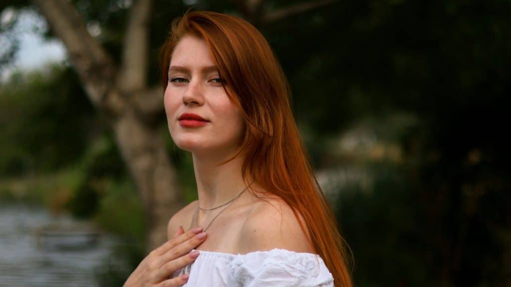 A woman with long red hair and a white top stands outdoors near a tree.