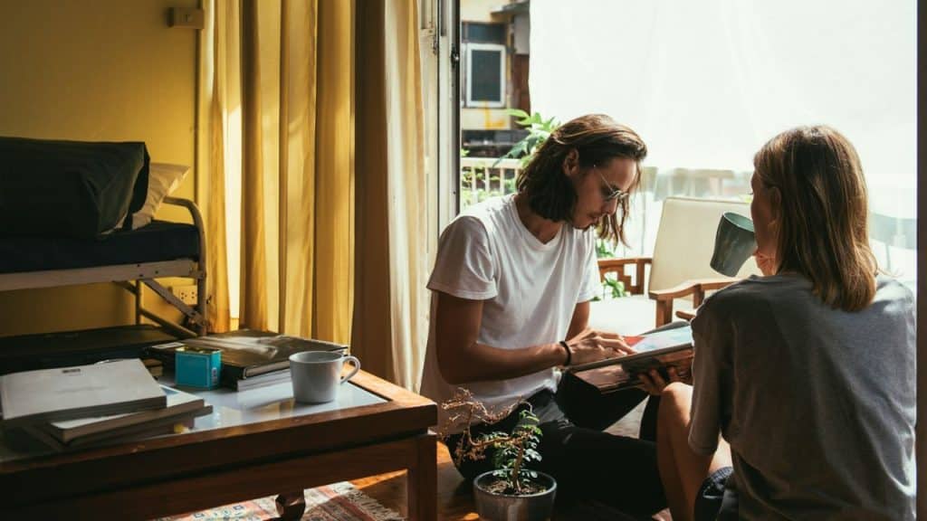 A man and woman sit on the floor of a sunlit room looking at a book.