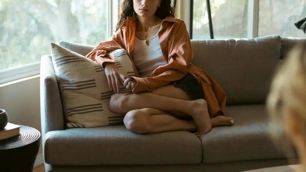A woman sitting cross-legged on a sofa in a sunlit living room, wearing a casual outfit.