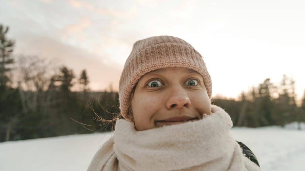 A person wearing a knit hat and scarf making a funny face outdoors at sunset.