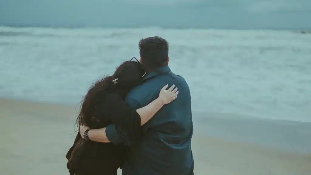 A rearview shot of a couple standing on a sandy beach, looking out at the ocean waves under an overcast sky.