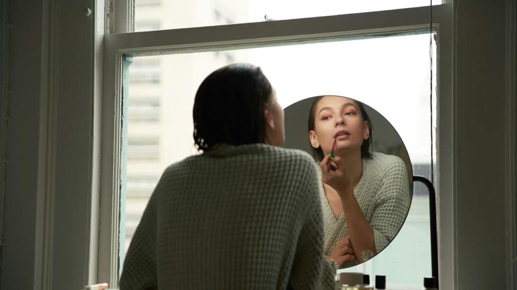 A woman applying lipstick while looking into a round mirror.
