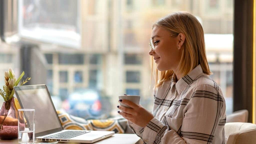 A woman in glasses smiles while holding a cup and sitting at a café table.