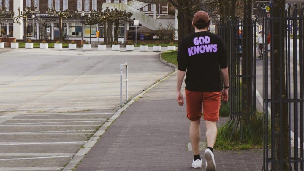 A man in a black shirt walks away on a sidewalk next to a parking lot.