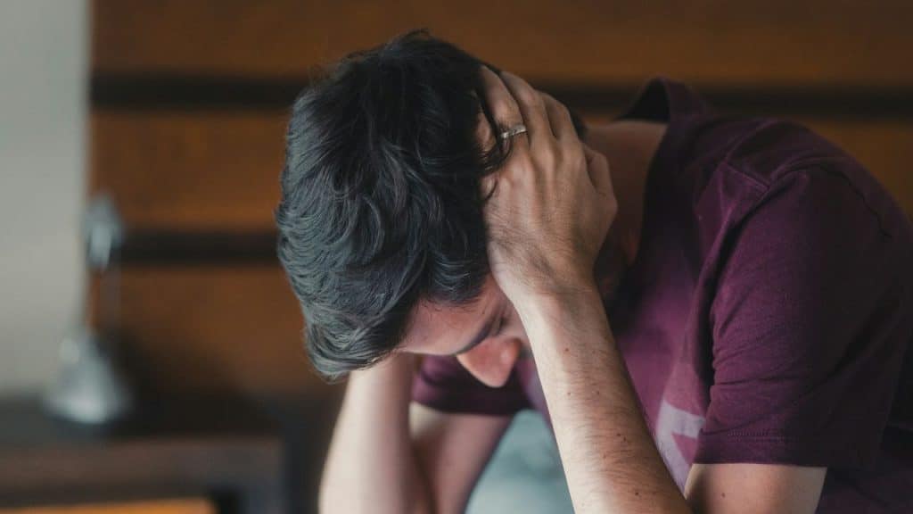 A man sits with his head down and his hands clasped behind his neck outdoors.