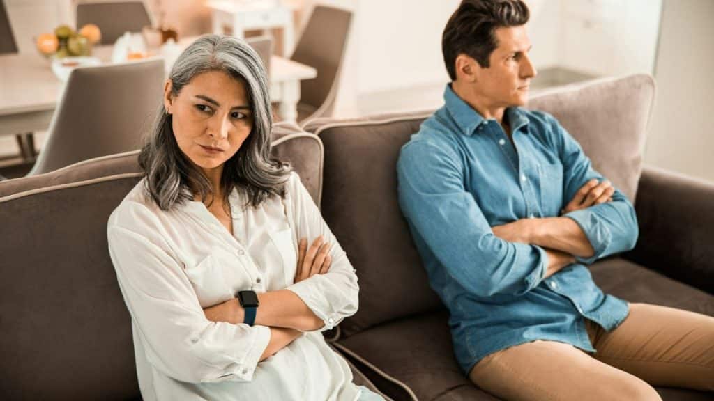 A man and woman sit on a sofa with their arms crossed, looking away.