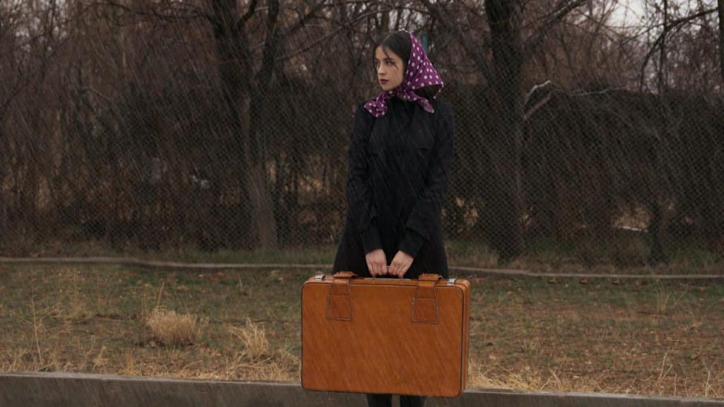 A woman standing on a road in the rain with a piece of luggage.