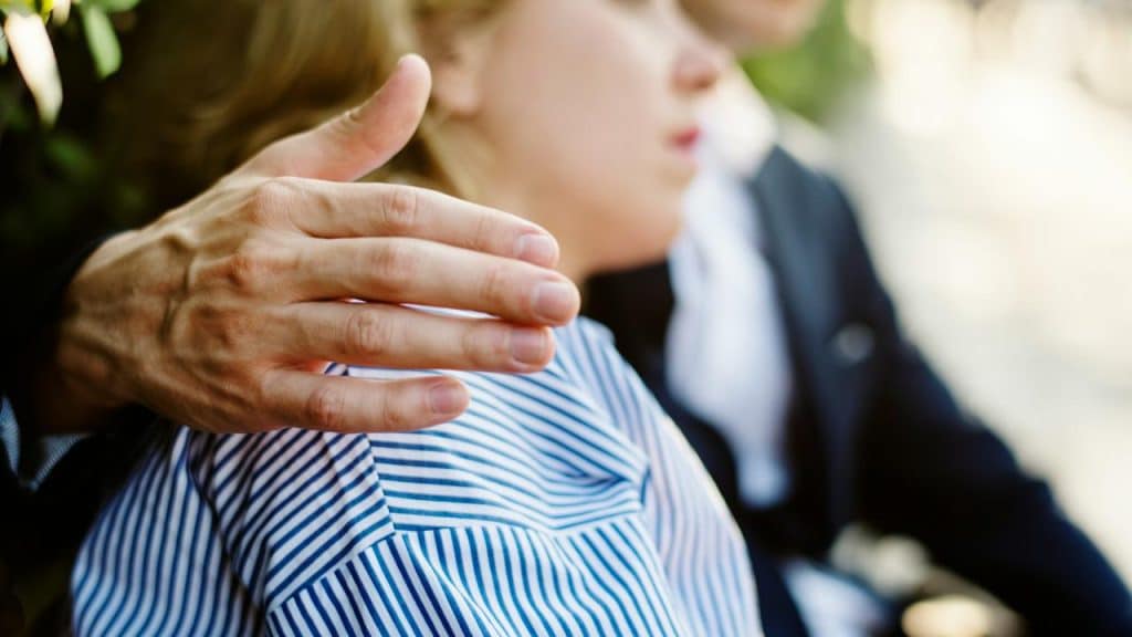 A man’s hand resting on a woman’s shoulder as they sit outdoors.