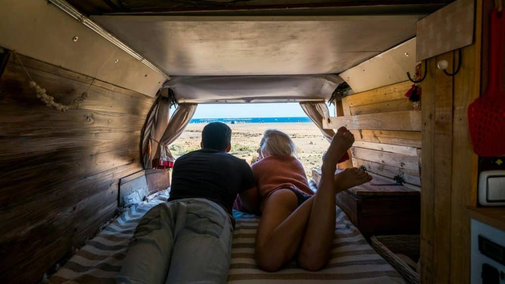A couple lies in the back of a van looking out at the beach.