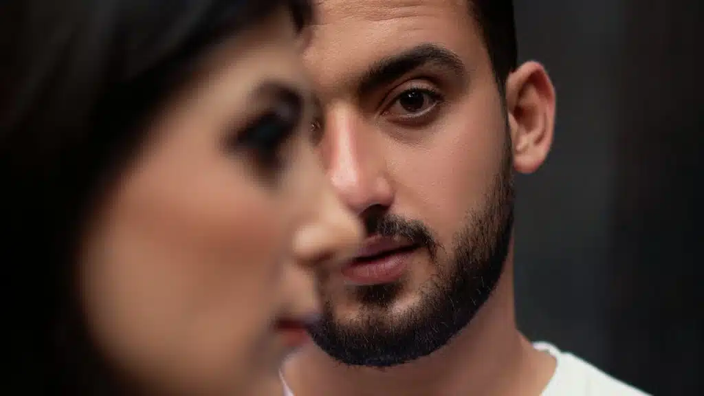 A close-up portrait of a bearded man in focus looking directly at the camera, partially obscured by the blurred profile of a woman in the foreground.