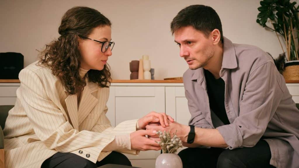 A woman in glasses holds a man's hands; both are looking serious, with a plant between them.