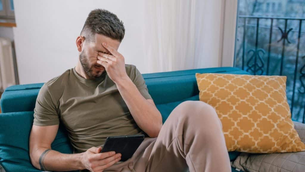 A man sits on a blue sofa, holding a tablet and pressing hand to face.