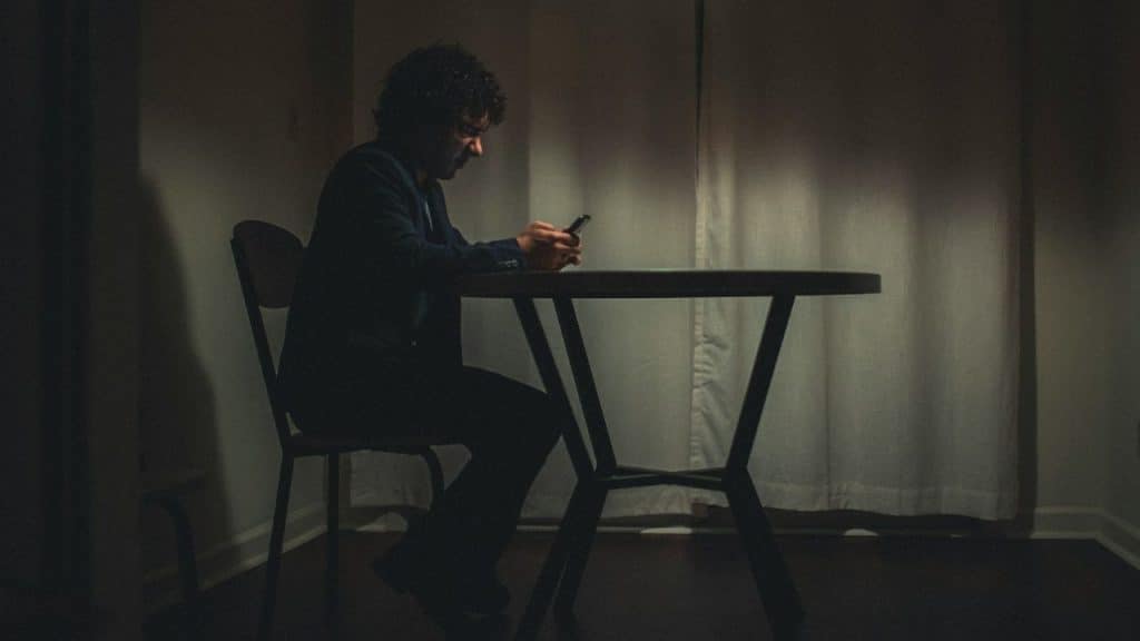 A man sits alone at a small round table in a dark room using a smartphone.