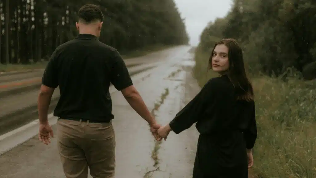 A man and woman walk away down a wet road while the woman looks back.